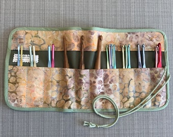 Puede incluir: Un estuche enrollable de tela verde con cierre de corbata marrón. El estuche tiene múltiples ranuras para guardar ganchos de ganchillo. Los ganchos son de varios colores, incluidos rojo, azul, verde y marrón.