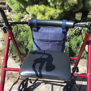 May include: A red walker with a black seat and a navy blue storage bag attached to the handlebars. The bag has a zippered pocket and is secured with blue straps. The walker is positioned outdoors with green foliage in the background.