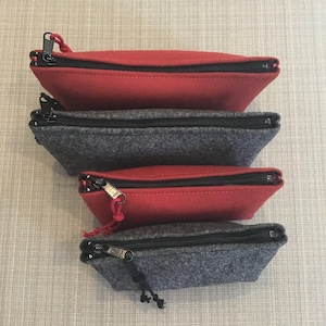 May include: Four felt pouches in red and gray, stacked. Each pouch has a black zipper and a matching pull cord. The pouches are trapezoidal in shape, with the red pouches on top and bottom, and the gray pouches in the middle.