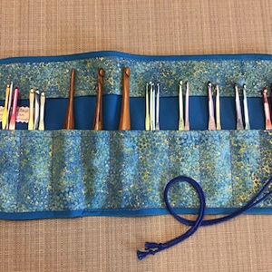 May include: A blue fabric crochet hook organizer with a batik-style pattern. The organizer holds various crochet hooks in different sizes and colors, including wooden and metal hooks. A blue cord secures the roll.