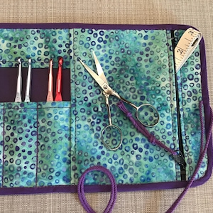 May include: A fabric roll-up organizer featuring a teal and blue circular pattern with purple trim. It contains six metal crochet hooks in gold, silver, and red, a pair of silver scissors, and a white measuring tape showing 2 inches.