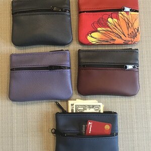 May include: Five small zippered pouches in different colors and patterns. The pouches are made of leather and have two zippered compartments. One pouch has a floral design in red, yellow, and orange. Another pouch has a blue floral design. The other pouches are black, purple, and burgundy.