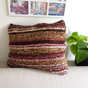 May include: A square throw pillow featuring a hand-knitted texture with horizontal stripes in shades of deep burgundy, warm brown, golden yellow, and cream. The yarn has a variegated appearance, creating a multi-tonal effect across the surface.