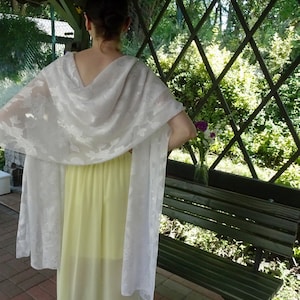Könnte beinhalten: Ein weißer Spitzenschal mit Blumenmuster, der über ein gelbes Kleid gelegt wird. Der Schal wird von einer Person getragen, die vor einem Holzgitter mit grünem Laub steht.