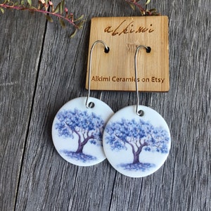 May include: Round white ceramic earrings featuring a blue tree design, suspended from silver hooks. The earrings are displayed on a wooden surface, with a wooden tag that reads "Alkimi Ceramics on Etsy".
