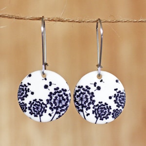 May include: A pair of white, circular drop earrings featuring a black dandelion motif. The earrings are suspended from silver-tone hooks. The dandelion design is detailed and artistic. The earrings hang against a neutral background.