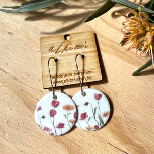 May include: A pair of white ceramic earrings with a floral pattern of red and orange flowers. The earrings are suspended from silver hooks. The earrings are displayed on a wooden tag with the text "alkimi" and "handmade ceramics www.alkimi.com.au".