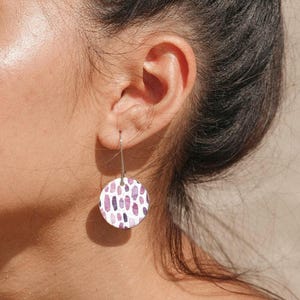 May include: A close-up of an ear with a circular earring. The earring has a white base with a pattern of purple and pink oblong shapes. It hangs from a silver hook. The earring is a piece of jewellery.