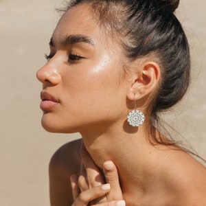 May include: A woman wearing a pair of round, silver earrings. Each earring has a white, circular design with a detailed, symmetrical pattern. The earrings hang from silver hooks.