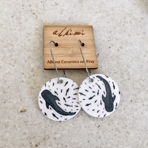 May include: Circular white earrings with a whale shark illustration in black and grey. The earrings hang from silver hooks. A wooden tag above displays the text "alkimi" and "Alkmi Ceramics on Etsy".