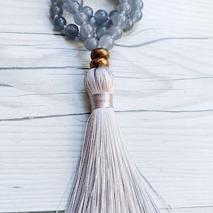 Op de afbeelding: Een grijze kralenketting met een kwast. De kralen zijn rond en hebben een lichtblauwe kleur. De kwast is gemaakt van een zacht, wit materiaal.