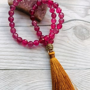May include: A pink gemstone mala with a gold tassel. The mala is made of round beads and is resting on a piece of amethyst.