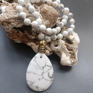 Peut inclure: Un collier de perles en howlite blanche avec un grand pendentif en howlite blanche. Le collier est sur une surface grise.