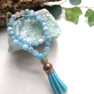 May include: A necklace made of light blue aquamarine beads. It has a gold-coloured metal accent and a light blue tassel. The beads are a mix of light blue and white, giving a soft, ocean-like look. The tassel is made of suede.