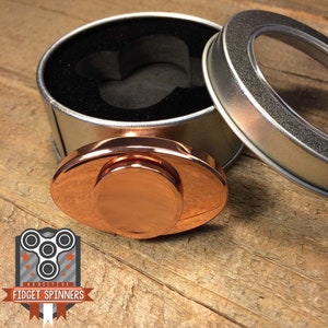 Puede incluir: Un spinner de fidget de color cobre en forma ovalada con un centro redondo. El spinner está en una caja de metal plateada con una tapa transparente.