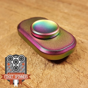May include: A metallic fidget spinner with a rainbow finish. The spinner has a rectangular body with rounded edges and a circular center piece. The text "ADDICTIVE FIDGET SPINNERS" is visible in the lower left corner.