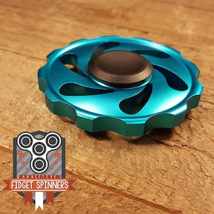 Könnte beinhalten: Ein türkisblauer Fidget Spinner mit einem braunen Mittelknopf liegt auf einer Holzoberfläche. Der Spinner hat ein zahnradähnliches Design mit geschwungenen Aussparungen. Das Logo "Addictive Fidget Spinners" ist unten links sichtbar.