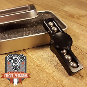 Könnte beinhalten: Schwarzer Fidget Spinner mit einem silbernen Metallzentrum und sechs silbernen Metallkugeln in einer Nut auf jeder Seite. Der Spinner befindet sich in einer silbernen Dose mit schwarzer Schaumstoffpolsterung.