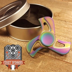 Puede incluir: Un spinner de fidget de metal iridiscente arcoíris con tres aspas y un rodamiento central. El spinner está sentado sobre una superficie de madera junto a una lata de plata con un interior de terciopelo negro.