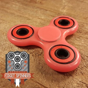 Puede incluir: Un spinner de mano rosa con tres anillos de rodamiento negros y rojos. El spinner tiene una tapa central rosa.