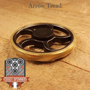 Puede incluir: Un spinner fidget negro y dorado con el texto "Arrow Tread" en la parte superior. El spinner tiene un centro negro y un anillo exterior dorado con un patrón de chevrón. El logotipo de Addictive Fidget Spinners está en la esquina inferior izquierda.