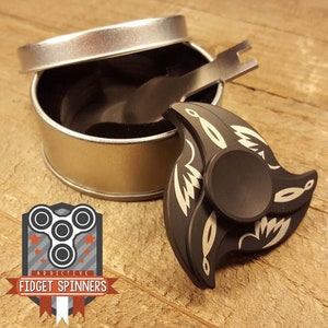 May include: Black metal fidget spinner with a silver design. The spinner is in a silver tin with a small tool for tightening the bearings.