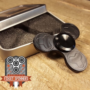 Puede incluir: Un spinner de fidget de metal negro con tres cuchillas redondeadas y un rodamiento central. El spinner está en una lata de plata con acolchado de espuma negra.