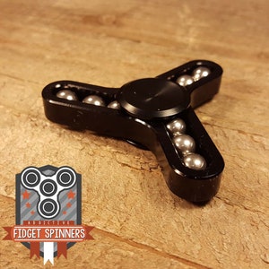Puede incluir: Fidget spinner de metal negro con un diseño de tres puntas. El spinner tiene un rodamiento central y está rodeado de rodamientos de bolas plateados.
