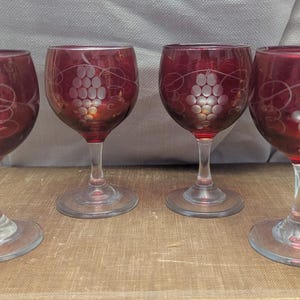 May include: Four ruby red wine glasses with etched grape cluster designs. The glasses have clear stems and bases, and are arranged on a wooden surface. The glasses are likely vintage and add a touch of elegance to any table setting.