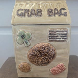 Peut inclure: Une jarre à biscuits en céramique en forme de sac en papier avec les mots "Grab Bag" imprimés sur le devant. La jarre est décorée d'images de biscuits, d'un trèfle et d'un motif rayé.