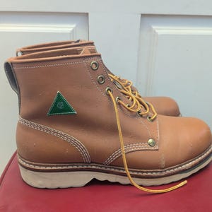 May include: Brown leather work boots with yellow laces. The boots feature a green triangle logo and white stitching. The soles are white. The boots are shown on a red surface.