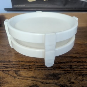 May include: A stack of two circular white plastic containers with a smooth finish. The containers feature vertical support ribs along the exterior sides that connect the two levels, creating a tiered structure with a flat top and open base design.
