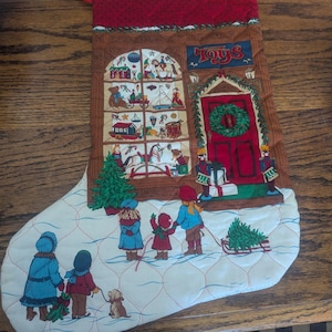 May include: A quilted Christmas stocking featuring a printed scene of children looking into a toy store window with a red door, a wreath, and toy soldiers. The lower section shows children in winter clothing walking in the snow with a small dog.