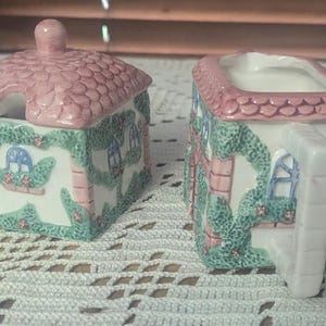 May include: A charming ceramic sugar bowl with a lid and a creamer, both shaped like quaint cottages. The roofs are pink, the walls are white with pink brick accents, and green vines with flowers adorn the sides. Windows are blue.