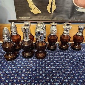 Op de afbeelding: Een verzameling van acht vintage schaakstuk-karaffen van bruin glas met zilverkleurige metalen stoppen in de vorm van schaakfiguren, waaronder koningen, koninginnen, paarden, torens en pionnen, opgesteld op een blauwe stof met bloemenpatroon.