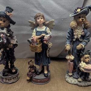 May include: Three decorative figurines. The first is a bear in a witch costume. The second is a girl with wings holding a basket. The third is a dog in a dress and hat. The figurines are on a brown surface.