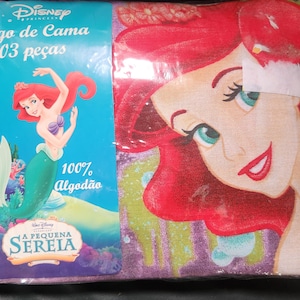 May include: A Disney Princess bedding set featuring Ariel from The Little Mermaid. The packaging shows Ariel with red hair and a green tail, with the text "Jogo de Cama 03 peças" and "100% Algodão". The bedding set has a colorful design.