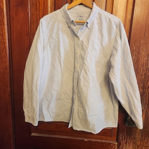 Op de afbeelding: Een lichtblauw en wit gestreept button-down overhemd met lange mouwen, hangend aan een houten hanger. Het overhemd heeft een borstzak en het merk "L.L. Bean" is zichtbaar op het label.