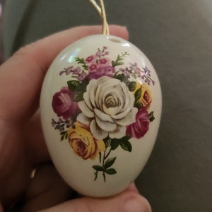 May include: A white ceramic egg-shaped ornament with a floral design. The ornament features a bouquet of pink, yellow, and white roses with green leaves.