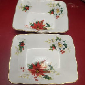 Könnte beinhalten: Zwei quadratische, weiße Keramikschalen mit Goldrand. Jede Schale hat ein weihnachtliches Blumendesign mit roten Weihnachtssternen, weißen Blumen, grünen Stechpalmenblättern und roten Beeren. Die Schalen dienen wahrscheinlich zum Servieren von Speisen.