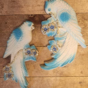 May include: Two ceramic bird wall hangings. The birds are white with blue accents on their wings and heads. They are perched on branches with blue and gold flowers. The background is a wooden surface.