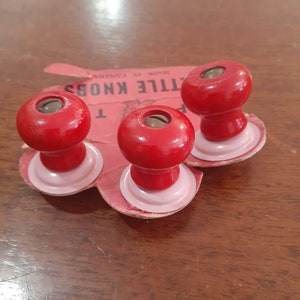 May include: Three red wooden knobs with a metal screw in the centre of each knob. The knobs are attached to a pink cardboard backing with the text "Little Knobs" and "Made in Canada".