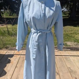 May include: A pale blue trench coat with a high, buttoned collar and a matching belt. The coat features long sleeves and decorative shoulder detailing. It is displayed on a mannequin. The coat is a classic style.