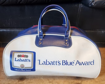 Pode incluir: Uma mala de viagem vintage branca e azul com o texto "Labatt's Blue Award" impresso na lateral. A mala tem uma parte superior e alças azuis, com um zíper vermelho. O logotipo da Labatt's é exibido em um quadrado azul.