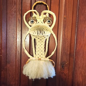 May include: A white macrame wall hanging with a whimsical frog design. The frog has large, round eyes with brown and white details. The macrame is made with a thick, white cord and has a fringe at the bottom.