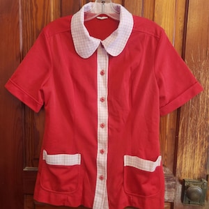 Peut inclure: Une chemise rouge vintage à manches courtes avec un col à carreaux blancs et roses, une fermeture boutonnée et deux poches avant. La chemise est suspendue à un cintre contre une porte en bois.