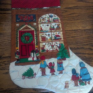 May include: A quilted Christmas stocking featuring a toy store storefront with the word TOYS above a red door. The scene includes children in winter clothing looking into a window display filled with toys, a small dog, and a snowy ground.