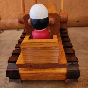 Puede incluir: Un bulldozer de juguete de madera con una figura roja, negra y blanca en el asiento del conductor. El bulldozer tiene un cuerpo de madera marrón y orugas negras.