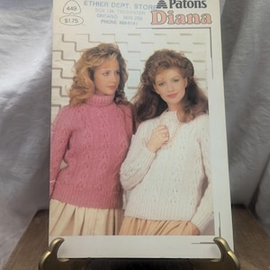 May include: A vintage magazine cover with two women wearing knitted sweaters. The cover reads "Patons Diana" and includes the price £1.38. The magazine is displayed on a gold-coloured stand.