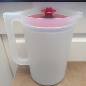 Può includere: Una brocca di plastica trasparente con coperchio e manico rossi. La brocca è smerigliata e il coperchio ha una piccola maniglia rotonda. La brocca è su una superficie di legno.
